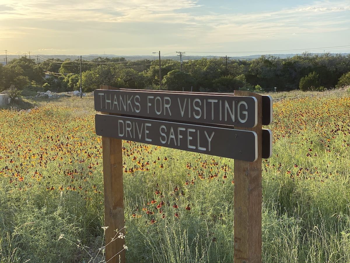 Carved brown sign reading Thanks for visiting, Drive safely in a wildflower field